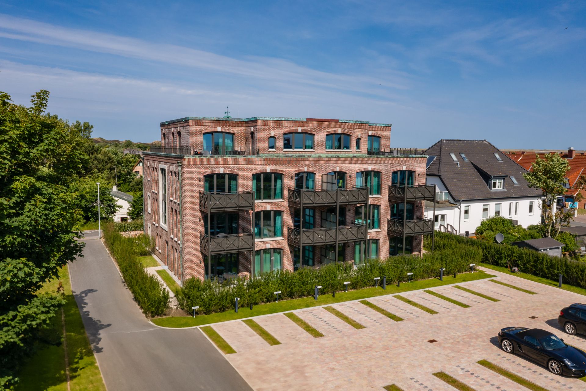 Mehrstöckiges Backsteingebäude mit Balkonen und angrenzendem Parkplatz, umgeben von Grün.