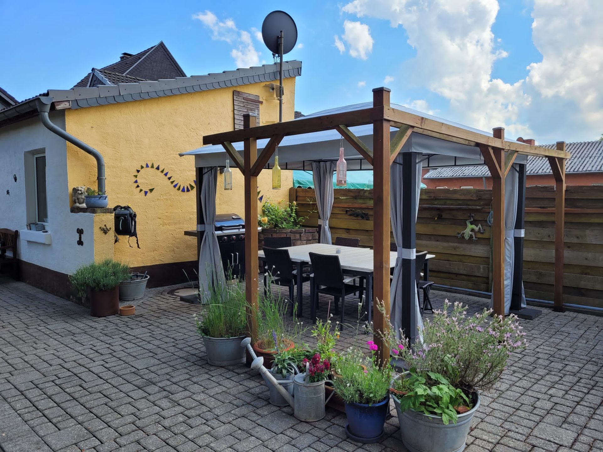 Überdachter Sitzbereich im Garten mit Tisch, Stühlen und Pflanzenkübeln auf Pflastersteinen.