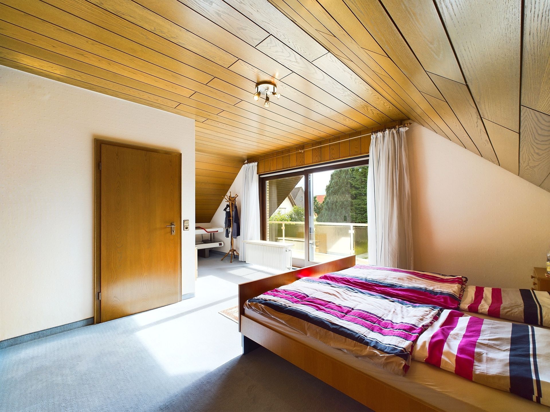 Helles Schlafzimmer mit Holzdecke, Doppelbett und Zugang zu einem Balkon mit Vorhängen.
