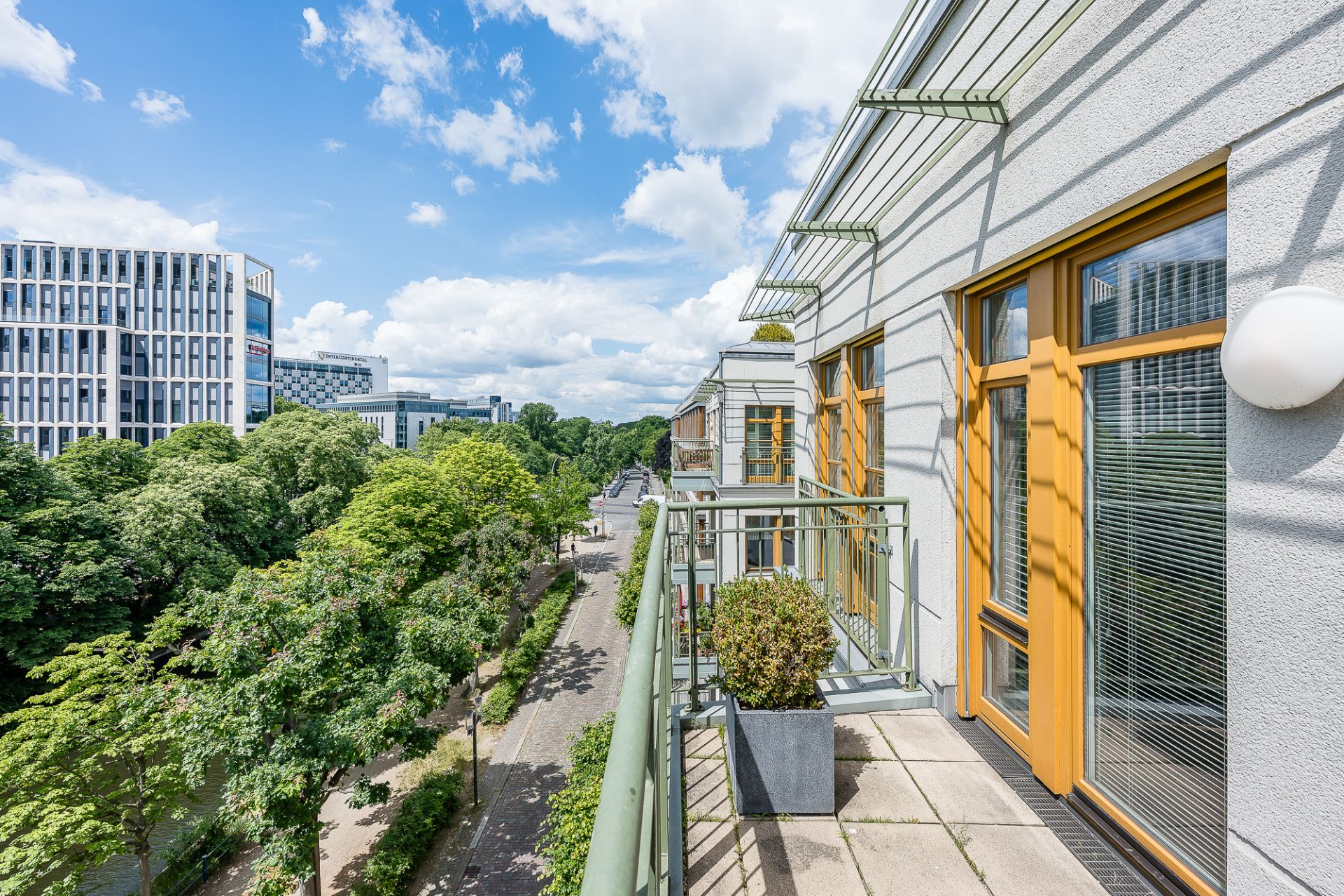 Balkon mit Pflanze, Blick auf Straße mit Bäumen und modernen Gebäuden bei blauem Himmel.