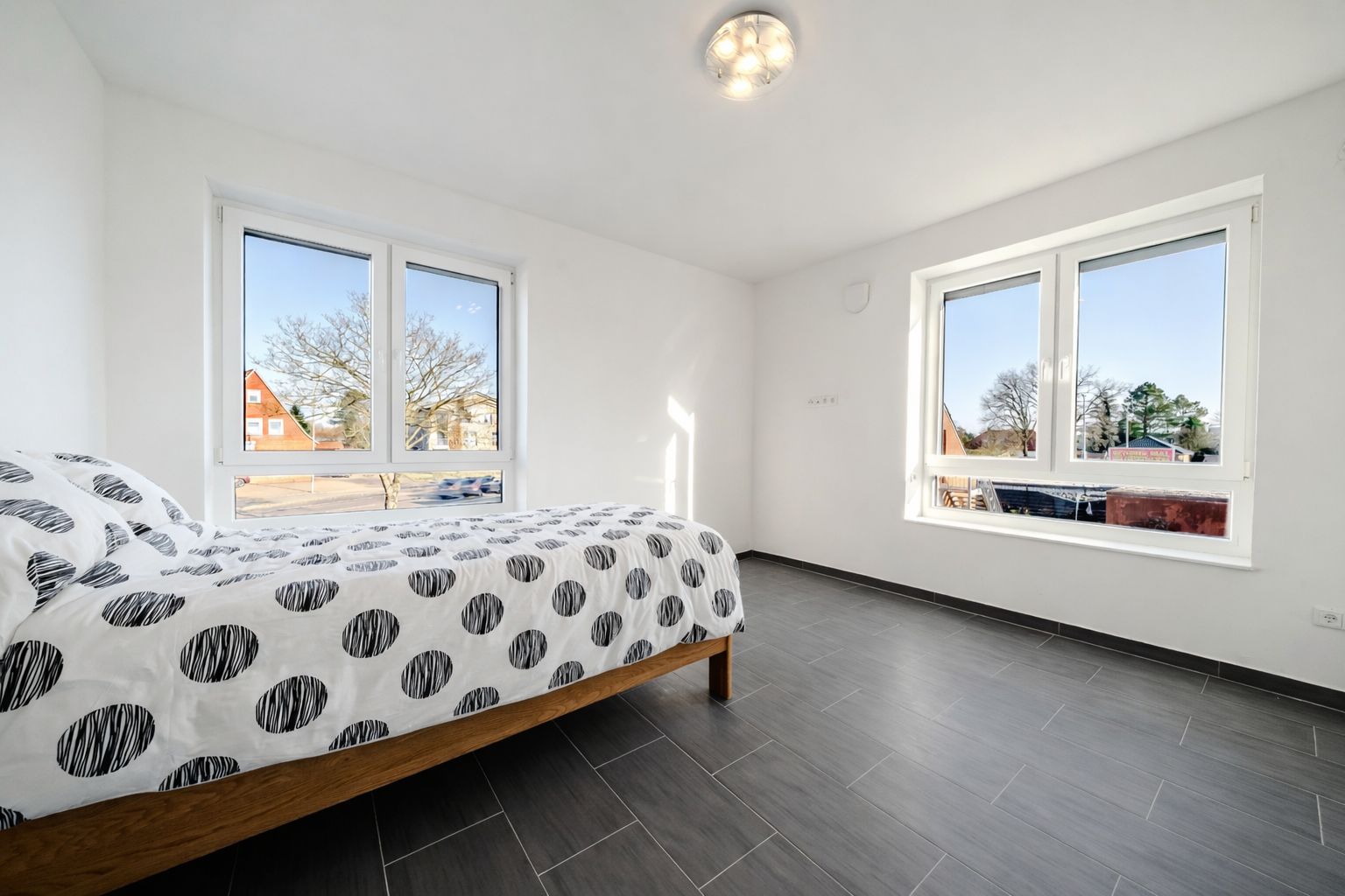 Helles Schlafzimmer mit Doppelbett, gemustertem Bettbezug und zwei großen Fenstern.