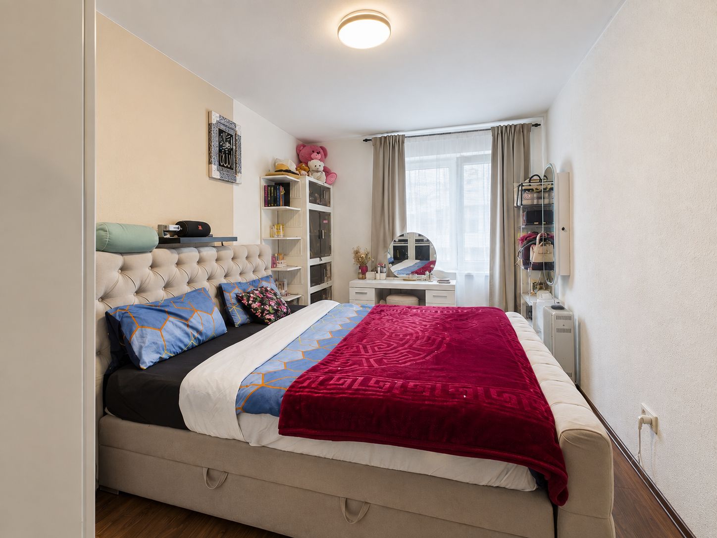 Schlafzimmer mit Doppelbett, Schminktisch, Regalen und Fenster mit Vorhängen.