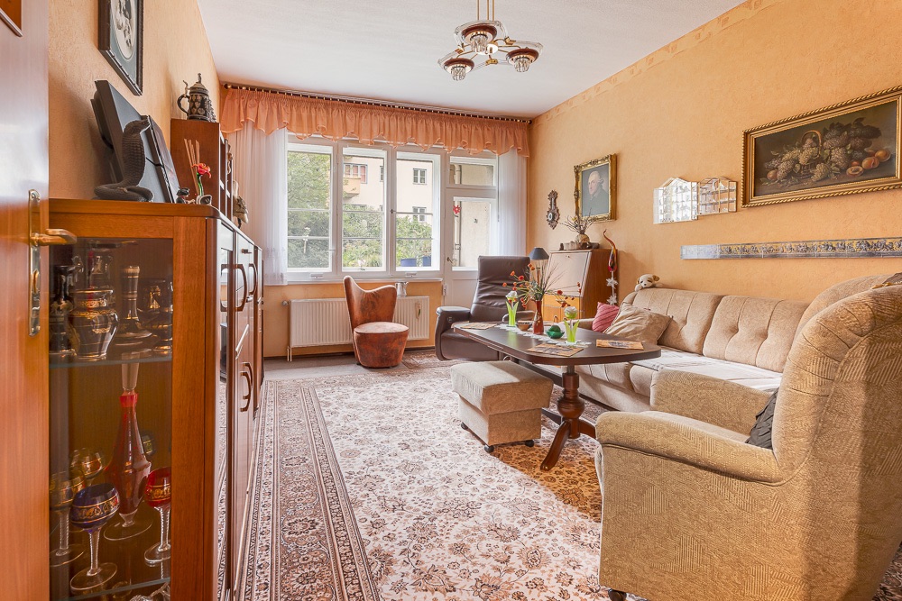 Wohnzimmer mit Sofa, Sessel, Teppich und Vitrine, große Fenster im Hintergrund.