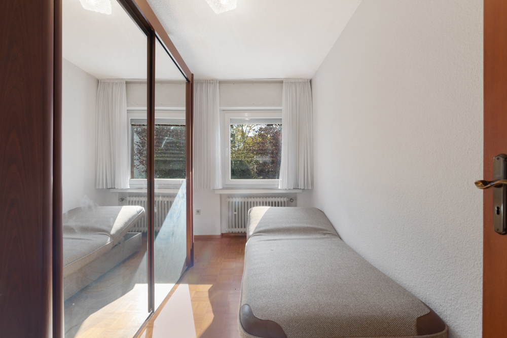 Kleines Schlafzimmer mit Einzelbett, großem Spiegel und zwei Fenstern mit weißen Vorhängen.
