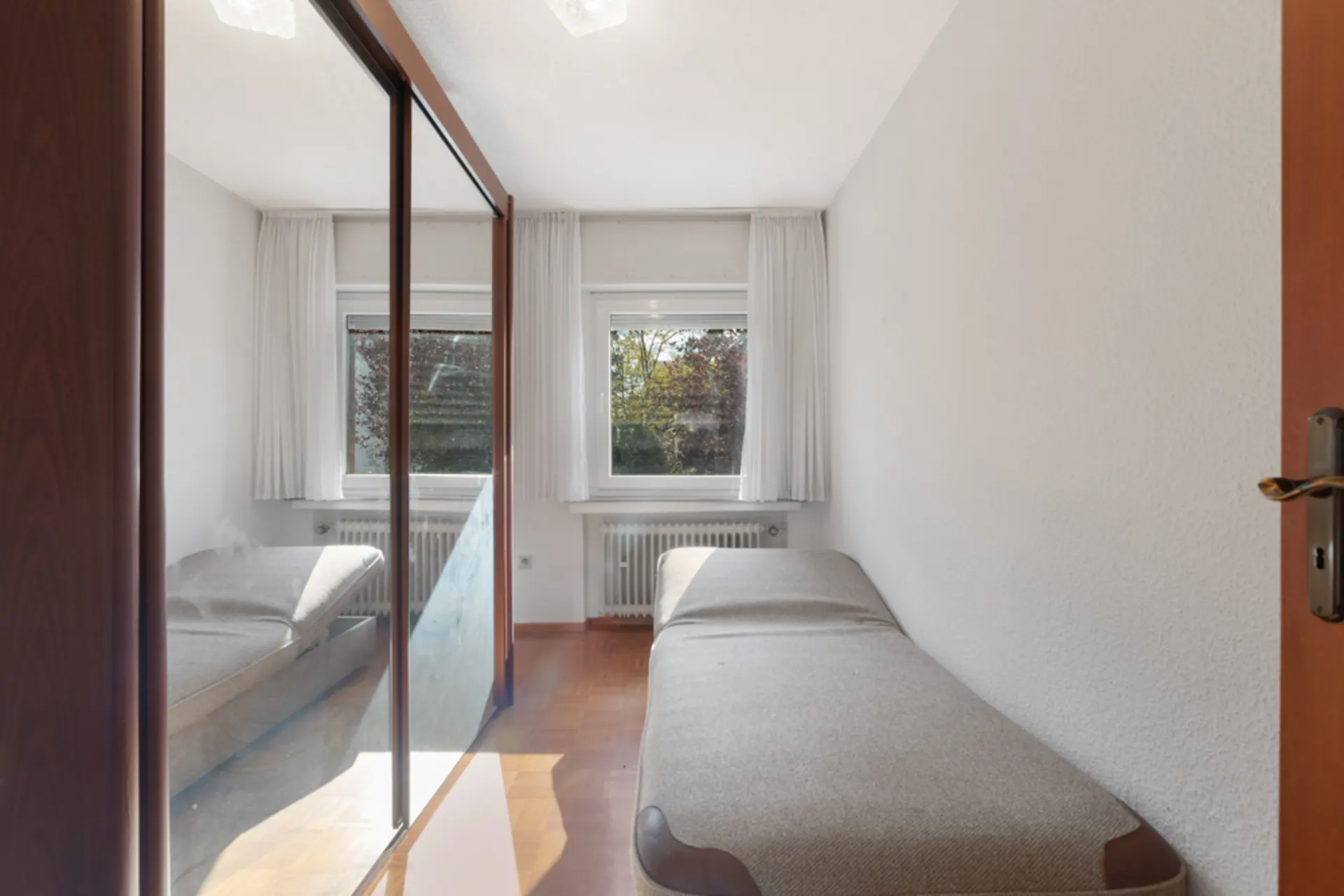 Kleines Schlafzimmer mit Einzelbett, großem Spiegel und zwei Fenstern mit weißen Vorhängen.