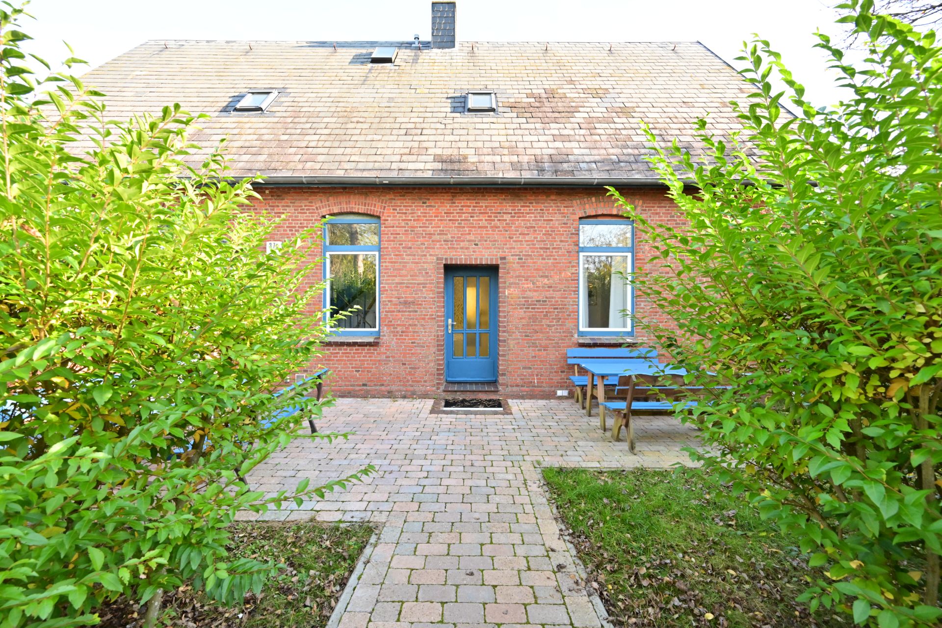 Backsteinfassade mit blauer Tür, zwei Fenstern und Sitzbank im Garten, umgeben von grünen Büschen.