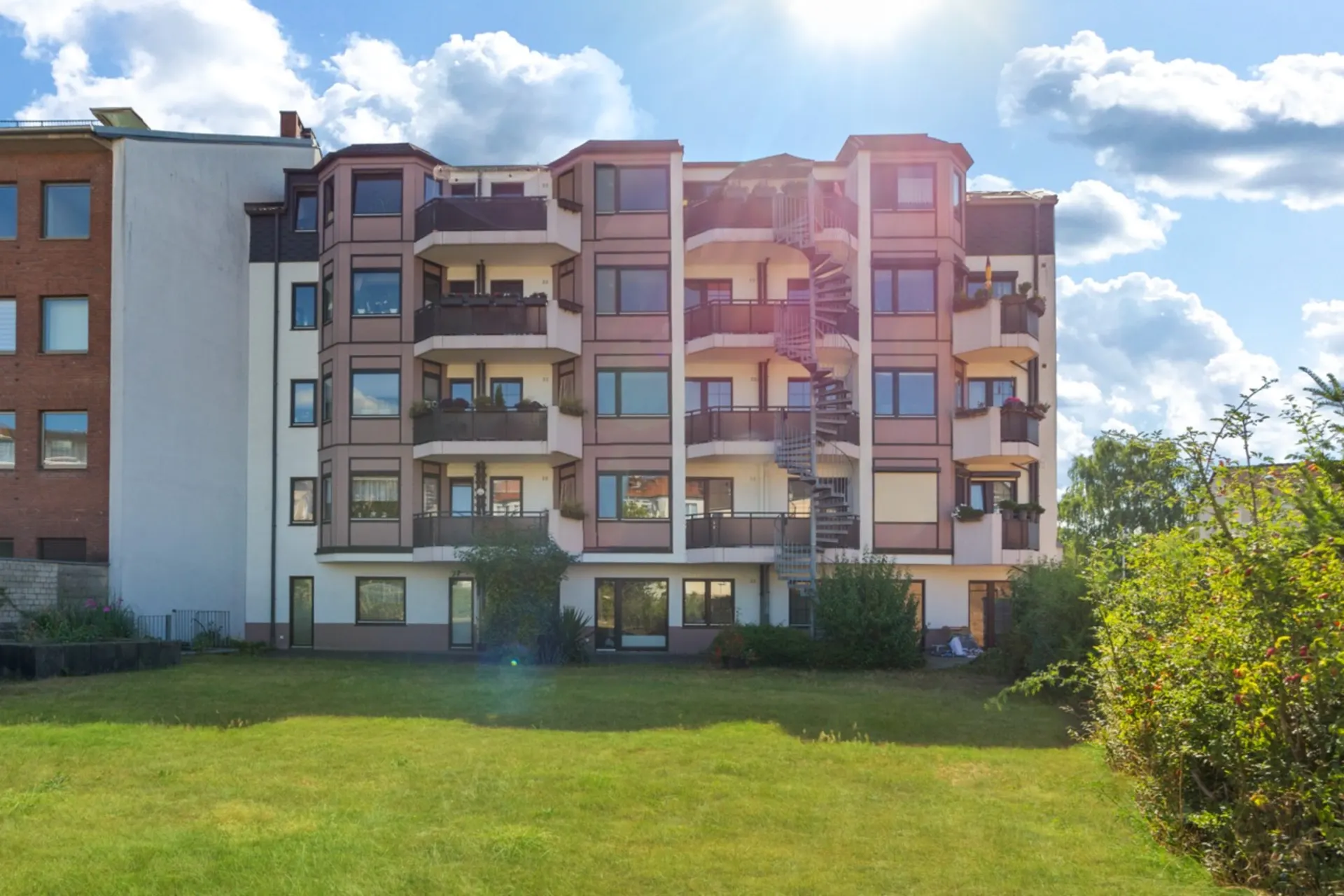 Mehrstöckiges Wohnhaus mit Balkonen, zentraler Wendeltreppe und Rasenfläche im Vordergrund.
