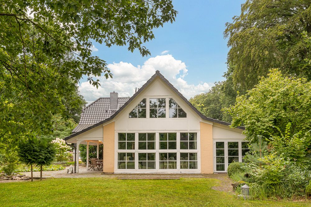 Modernes Haus mit großen Fenstern, Satteldach und Garten, umgeben von Bäumen und Rasen.