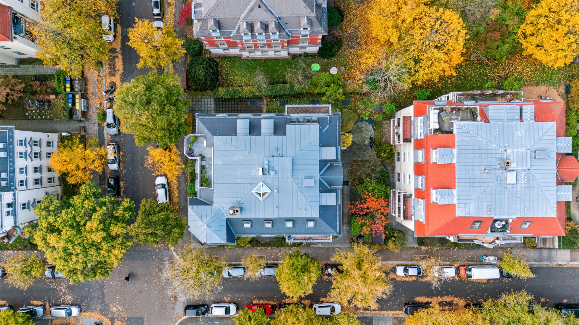 Luftaufnahme von Wohnhäusern, Bäumen und parkenden Autos an einer Straße im Herbst.