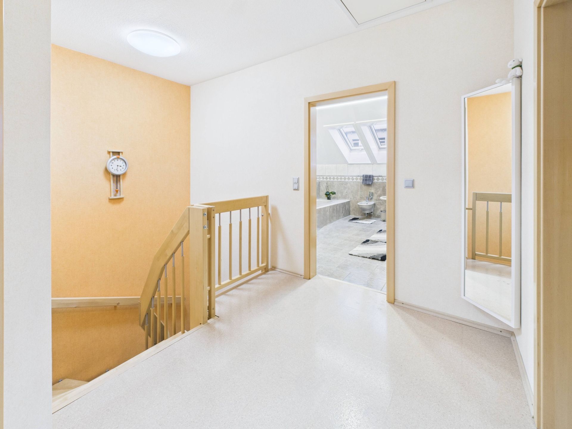 Heller Flur mit Treppe, Wanduhr und Blick in ein Badezimmer mit Dachfenstern.