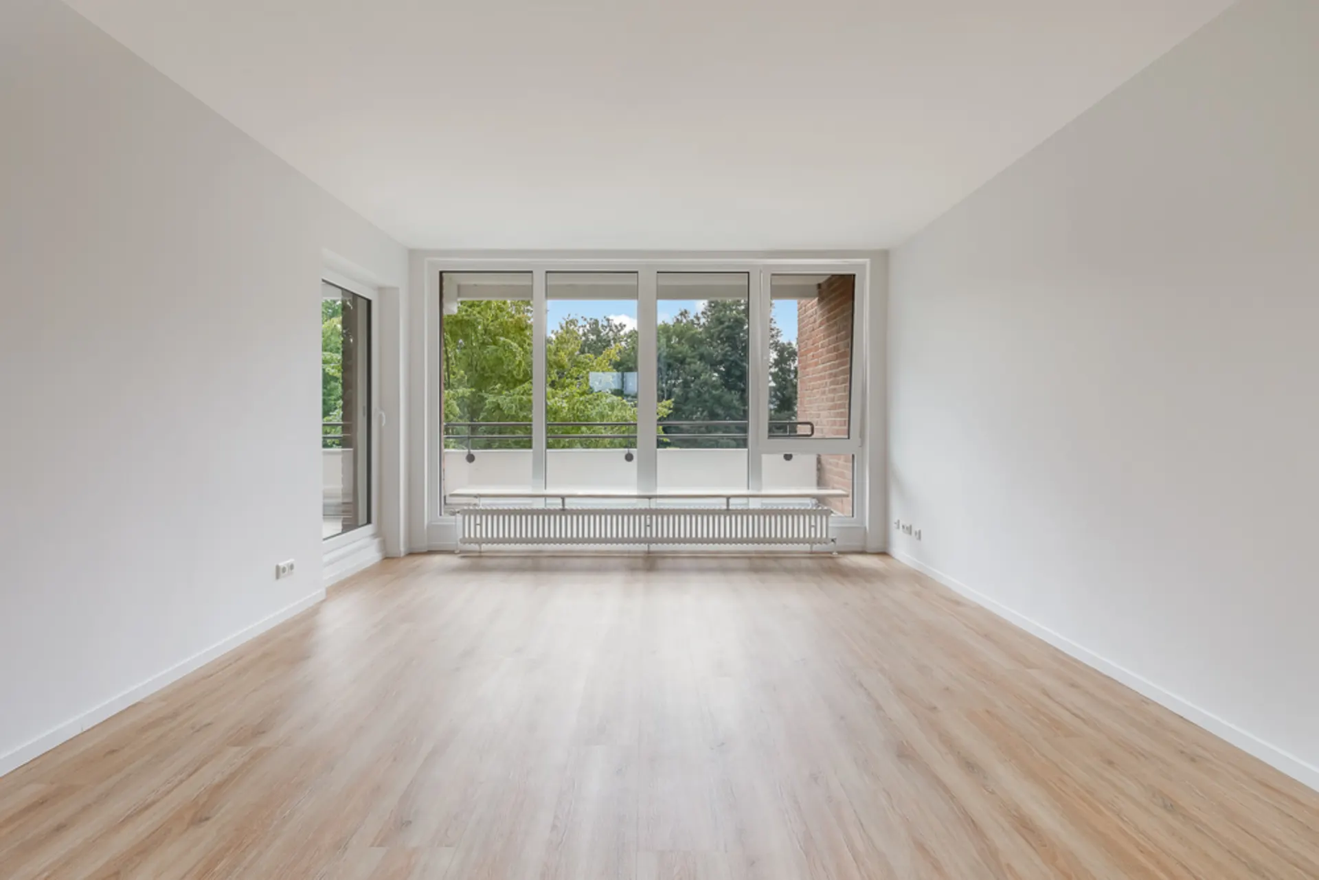 Leerer, heller Raum mit Holzboden und großen Fenstern mit Blick ins Grüne.