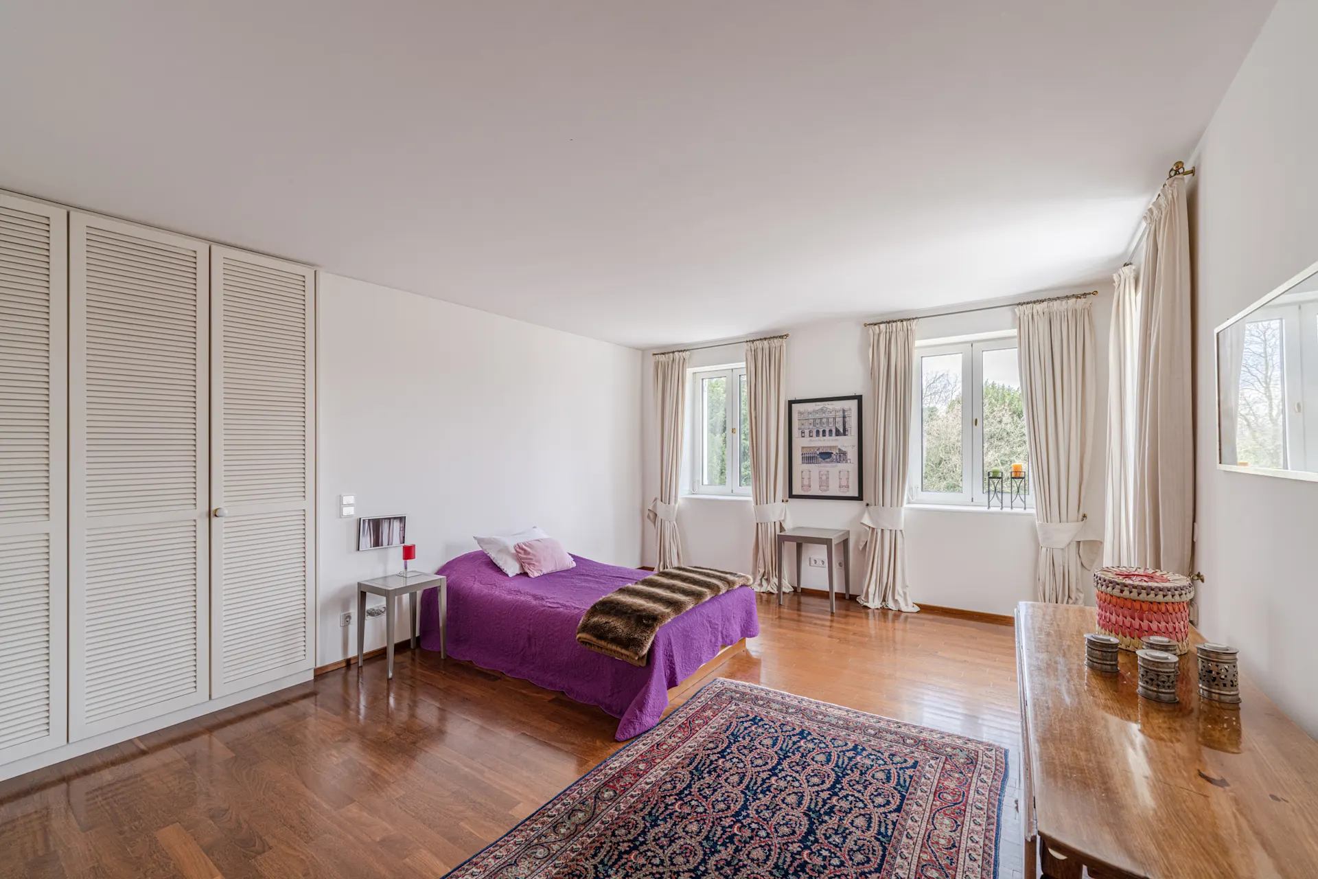 Helles Schlafzimmer mit Einzelbett, Holzfußboden, Teppich und großen Fenstern mit Vorhängen.