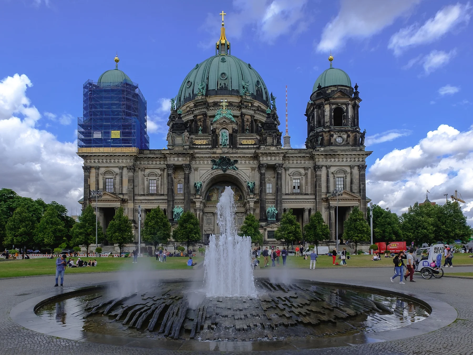 Großer Brunnen mit Wasserstrahl vor einem historischen Gebäude mit grüner Kuppel und Gerüst.