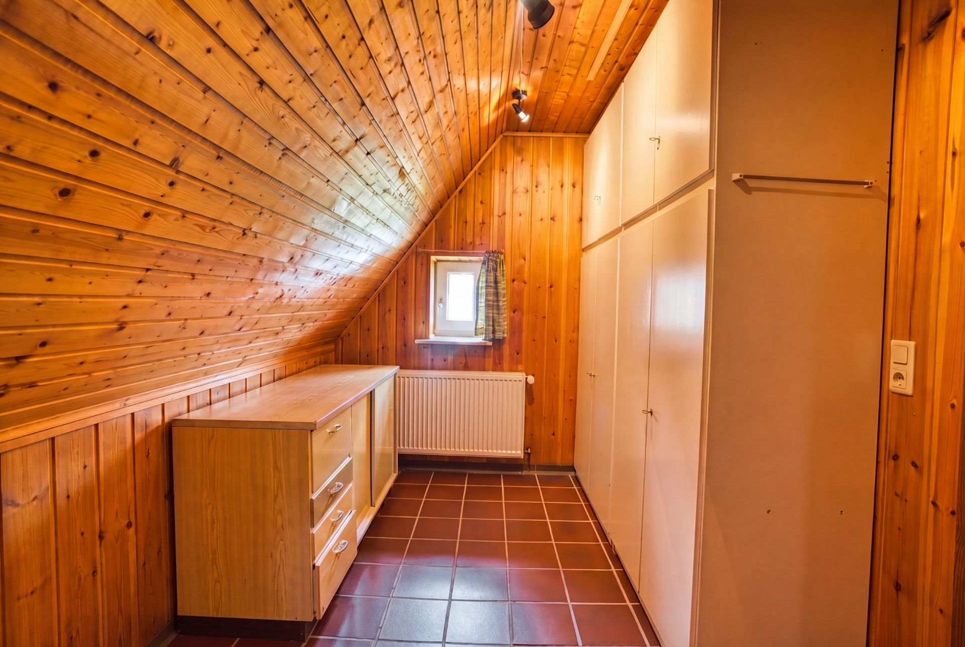 Schmaler Raum mit Holzverkleidung, Fliesenboden, Schränken und kleinem Fenster am Ende.