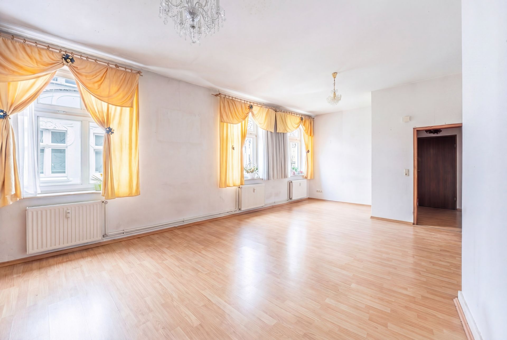 Heller, leerer Raum mit Holzboden, großen Fenstern und gelben Vorhängen.