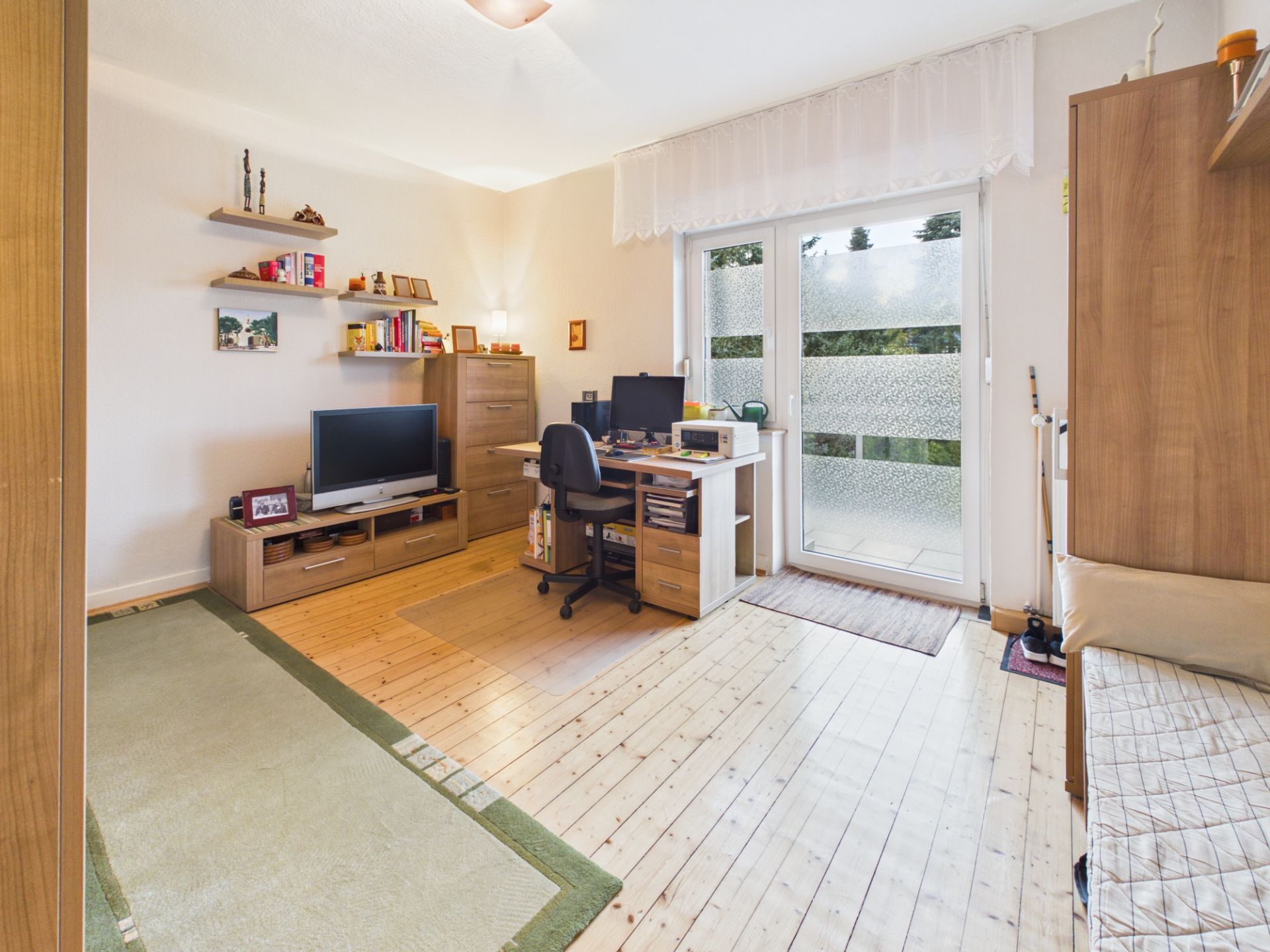Wohnzimmer mit Schreibtisch, Fernseher, Regalen und Zugang zu einem Balkon mit Glastür.