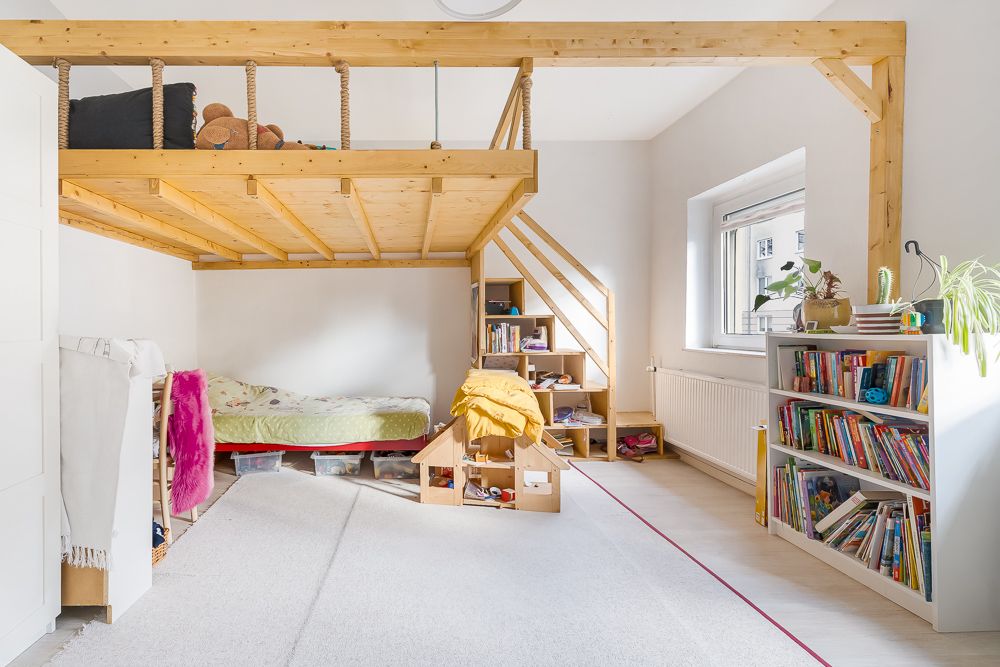 Helles Kinderzimmer mit Hochbett aus Holz, Bücherregalen, Fenster und Teppichboden.