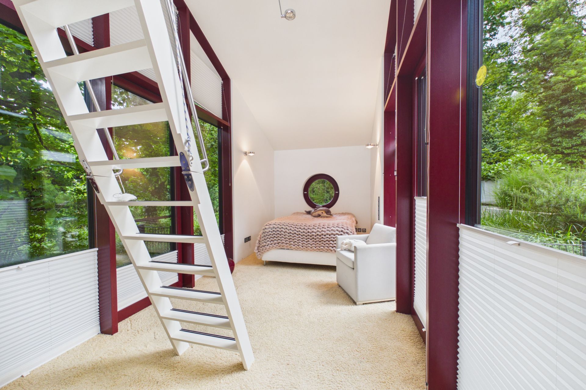 Helles Schlafzimmer mit Teppich, großem Fenster, Bett, Sessel und weißer Treppe nach oben.