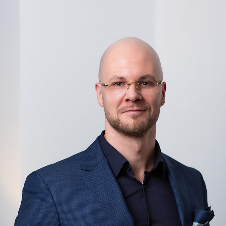 Mann mit Brille und Bart trägt blauen Anzug und steht vor weißer Wand.