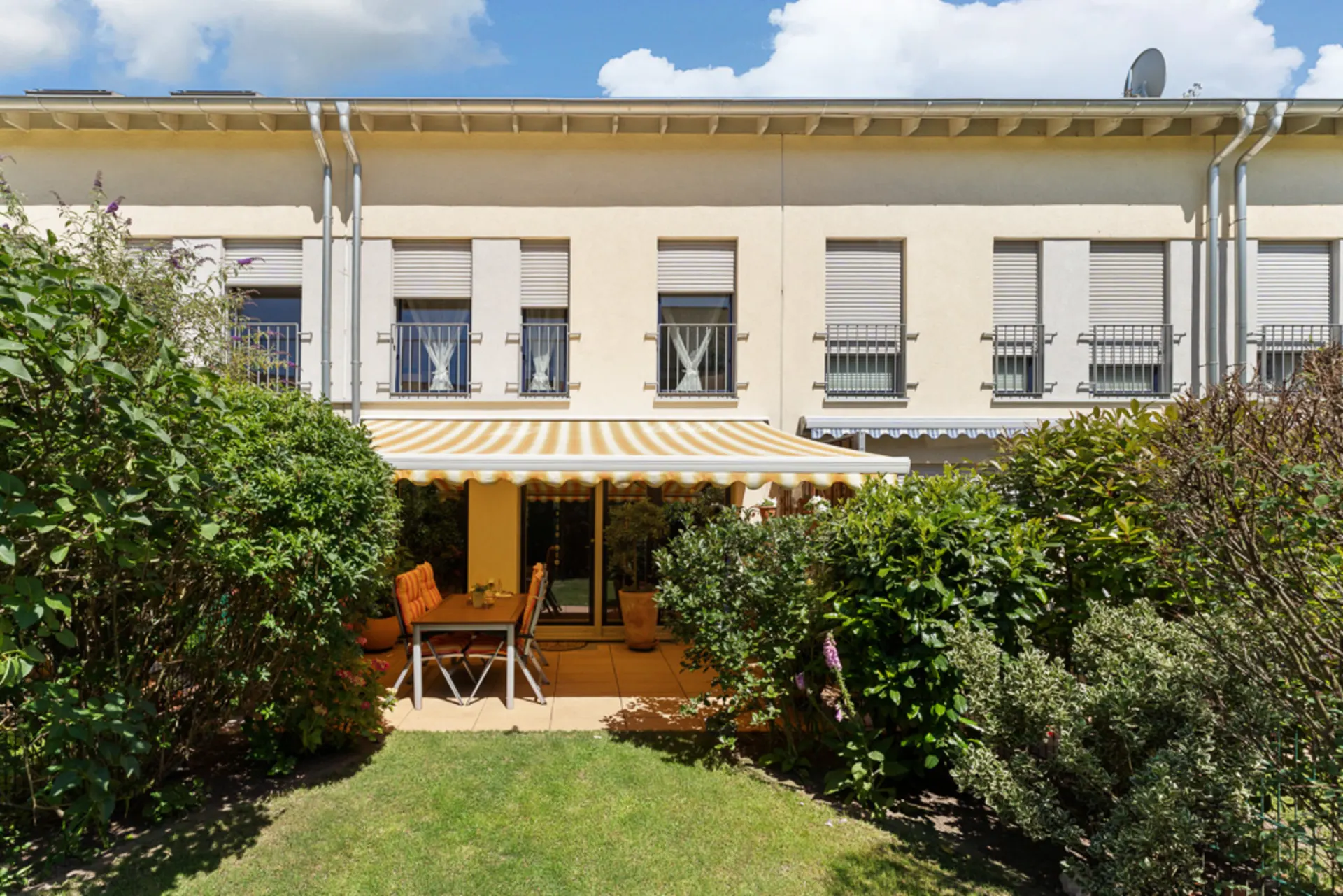 Terrasse mit Markise, Tisch und Stühlen vor einem Wohnhaus, umgeben von Garten und Sträuchern.