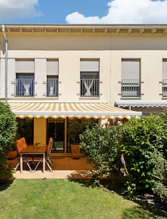 Terrasse mit Markise, Tisch und Stühlen vor einem Wohnhaus, umgeben von Garten und Sträuchern.