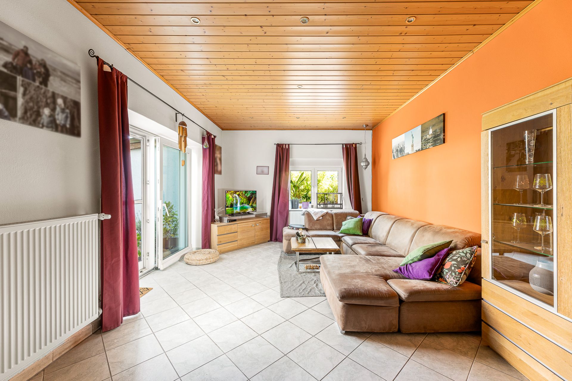 Helles Wohnzimmer mit braunem Sofa, Glastür, Fernseher und oranger Wand.
