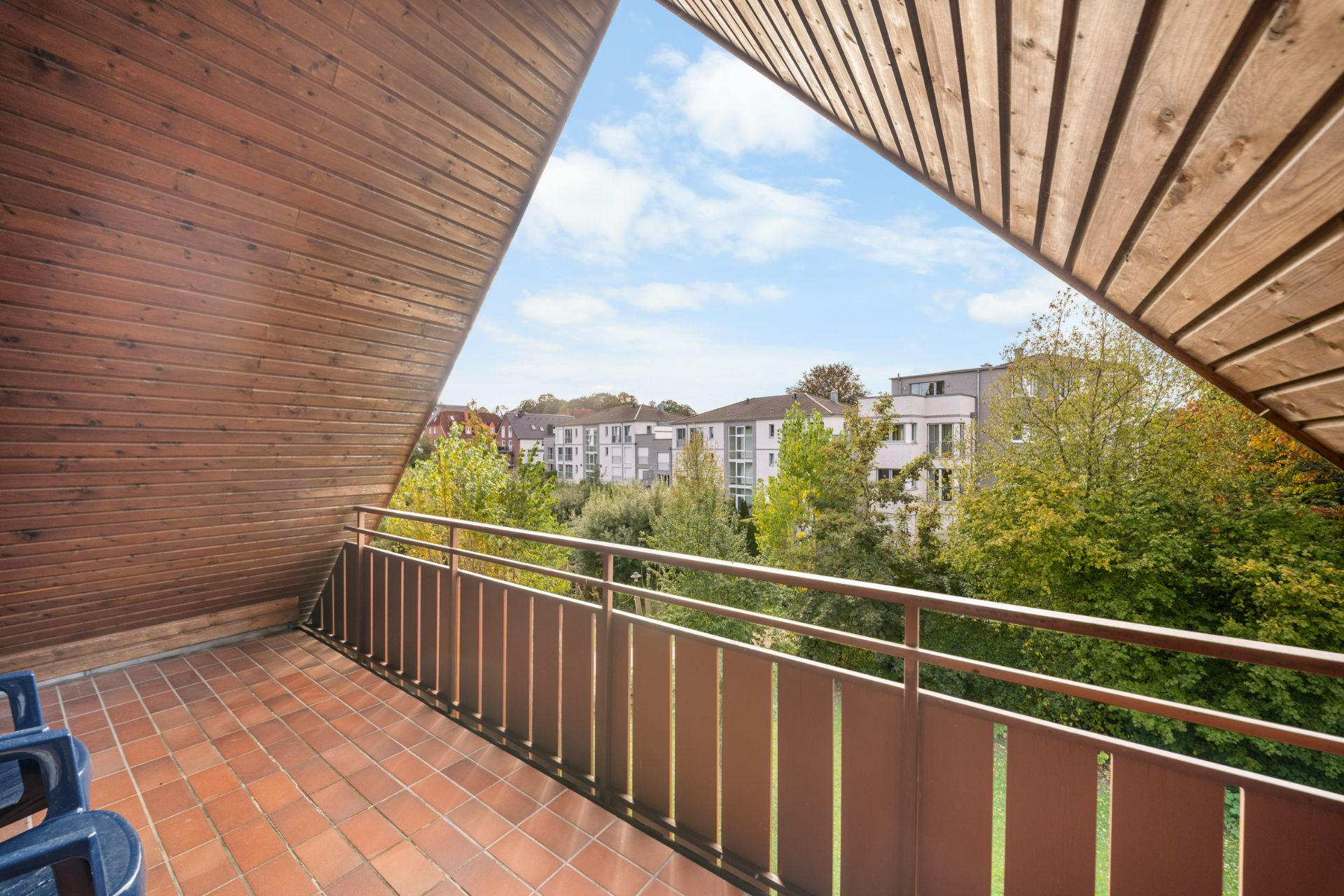 Überdachter Balkon mit Holzwand, Blick auf Bäume und benachbarte Wohngebäude.