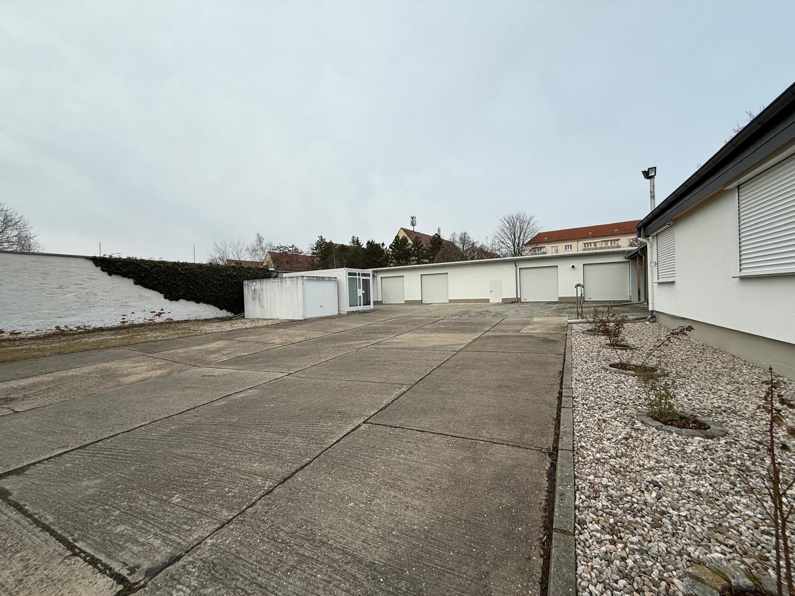 Großer betonierter Hof mit mehreren Garagen und weißem Wohnhaus, wenig Bepflanzung.