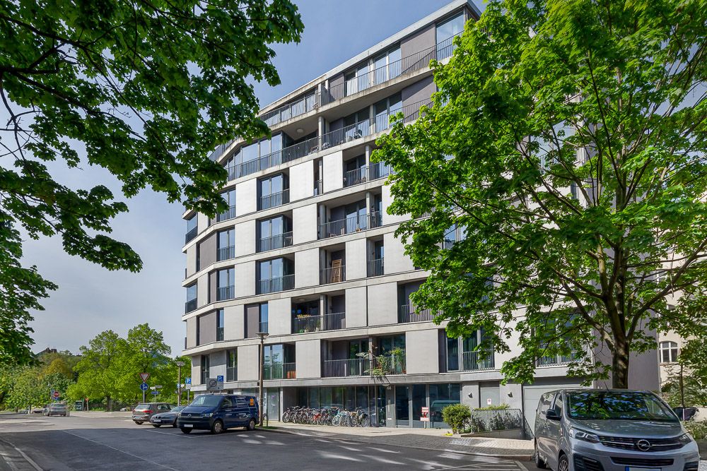 Modernes Wohngebäude mit Balkonen, Bäumen und geparkten Autos an der Straße.