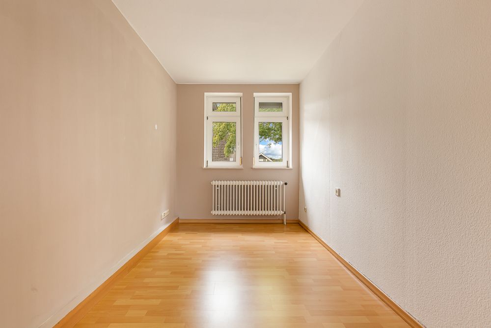 Leerer Raum mit Holzfußboden, zwei Fenstern und einem Heizkörper unter den Fenstern.