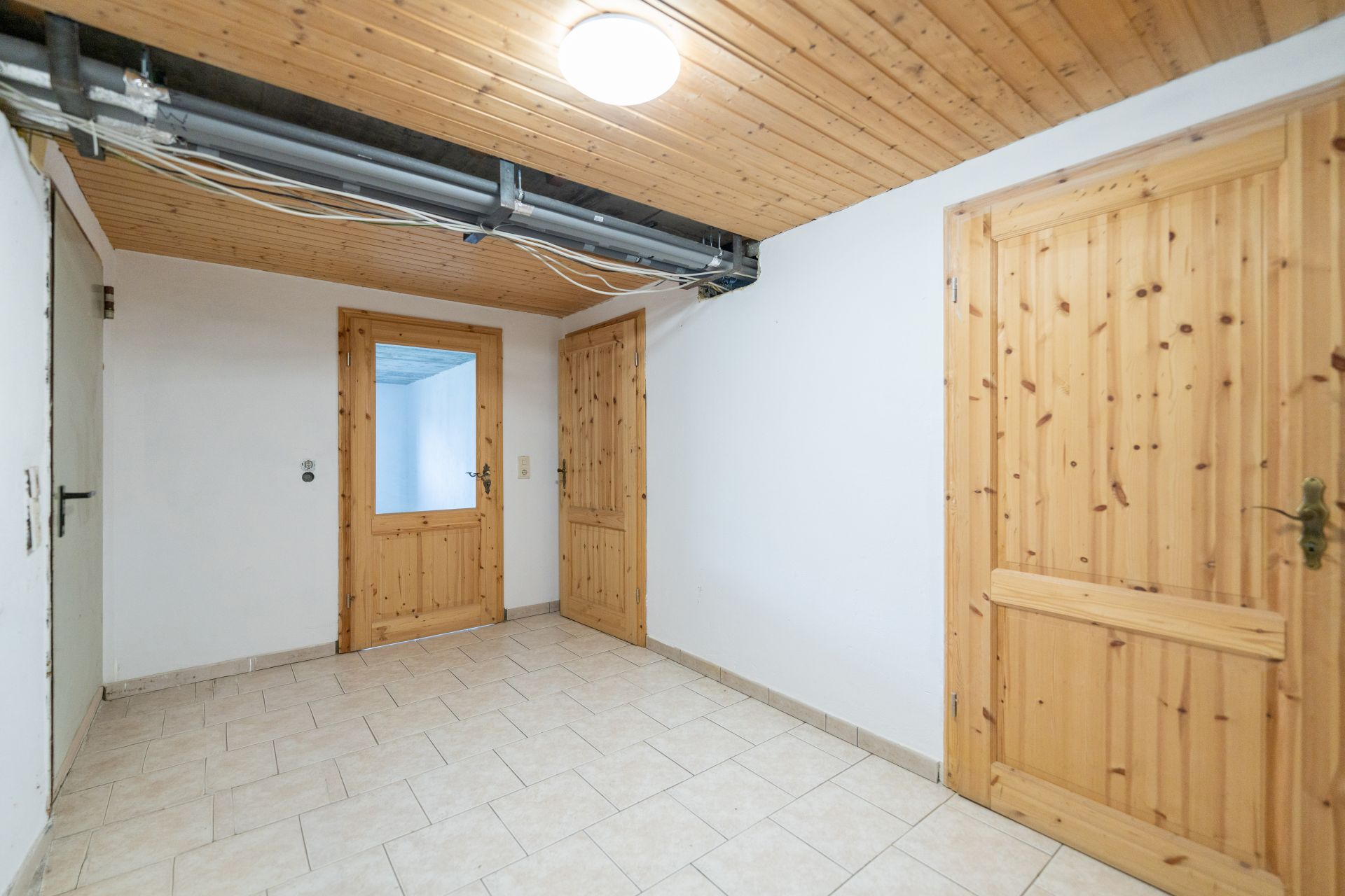 Heller Flur mit drei Holztüren, Fliesenboden und Holzdecke mit sichtbaren Leitungen.