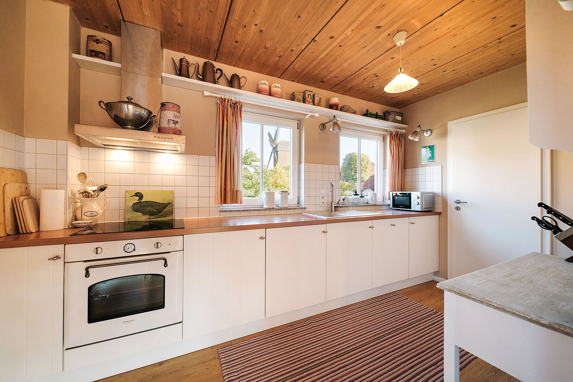 Helle Küche mit Holzdecke, weißen Schränken, zwei Fenstern und Küchengeräten.