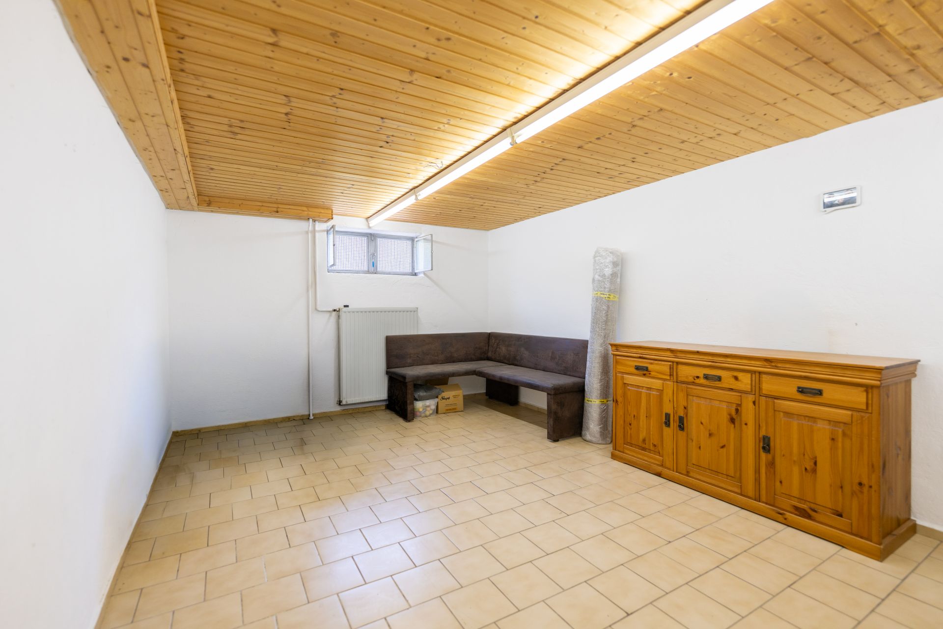 Kellerraum mit Holzmöbeln, Ecksofa, Kommode und kleinem Fenster unter Holzdecke.