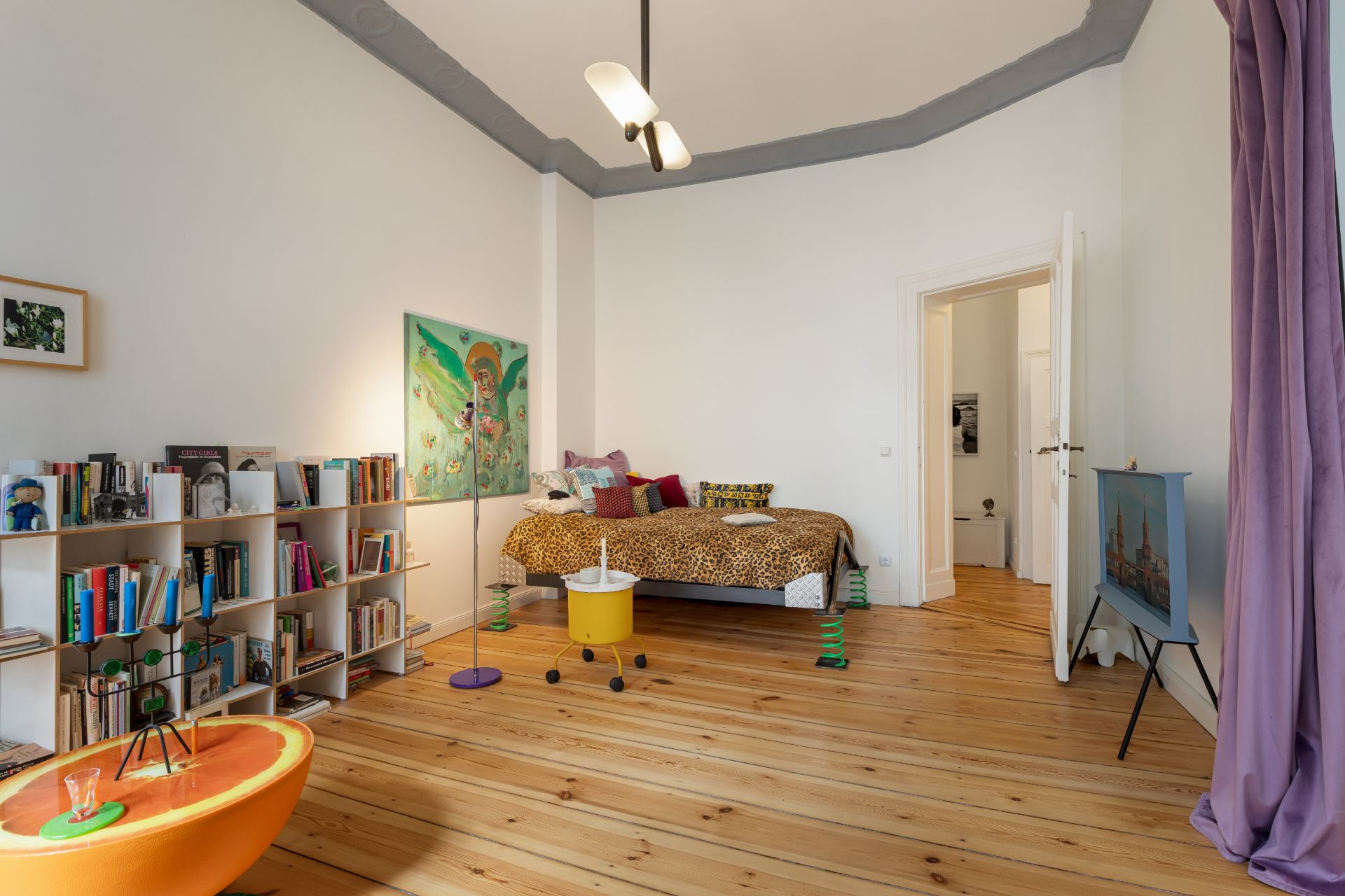 Helles Schlafzimmer mit Holzboden, Bett, Bücherregal und bunten Dekorationen.