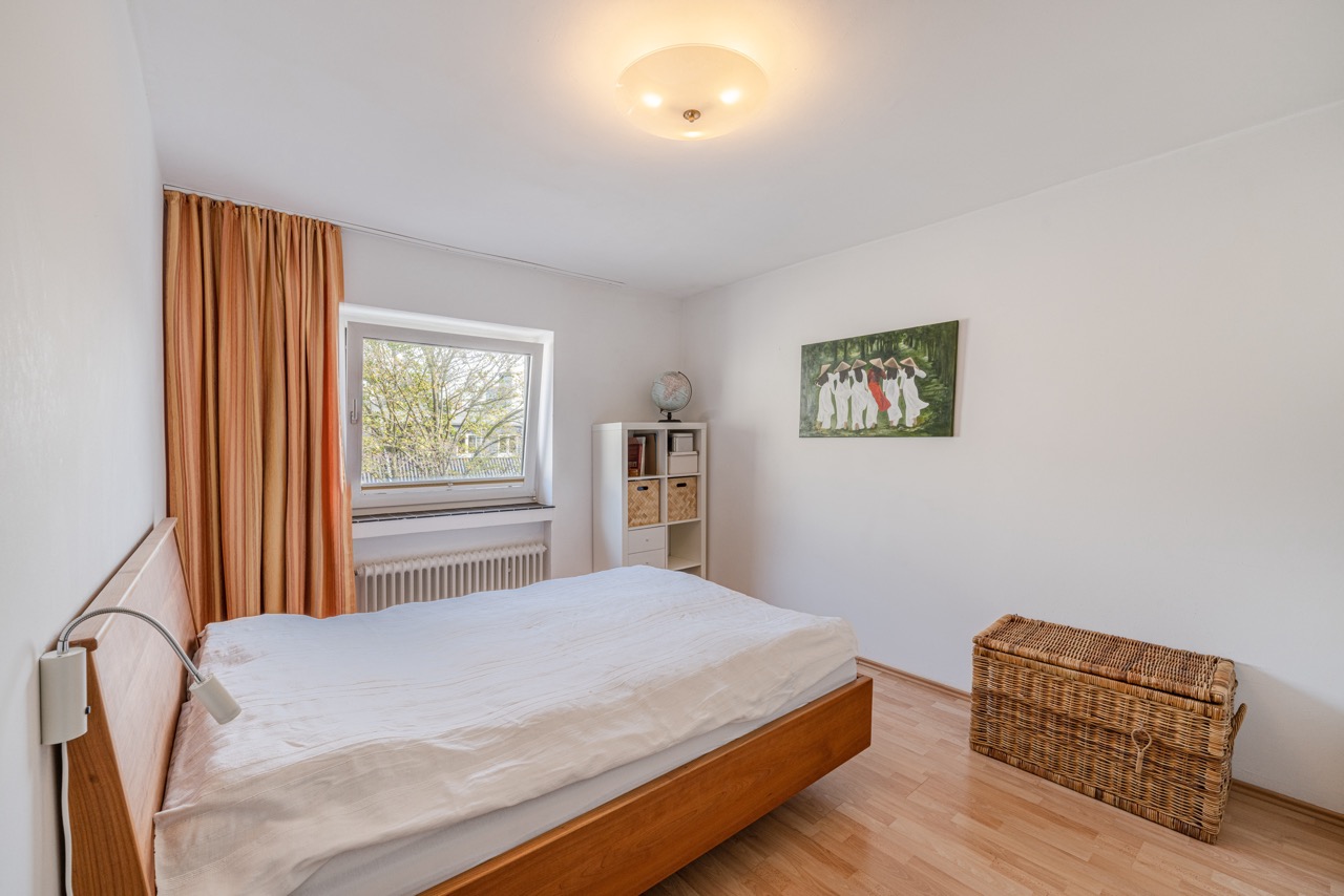 Helles Schlafzimmer mit Holzbett, Fenster, Regal und Korbtruhe auf Holzfußboden.
