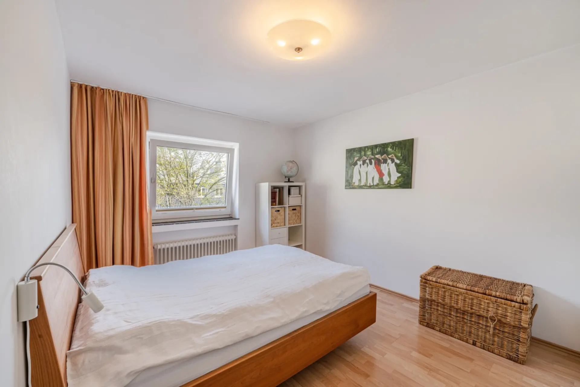 Helles Schlafzimmer mit Holzbett, Fenster, Regal und Korbtruhe auf Holzfußboden.