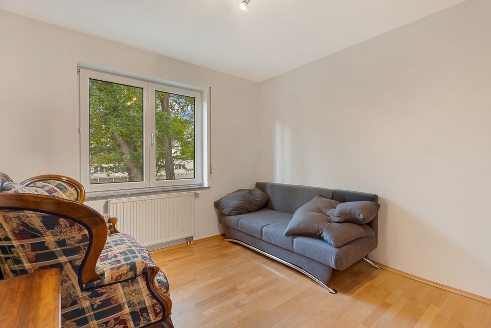 Helles Wohnzimmer mit grauem Sofa, Sessel und großem Fenster mit Blick auf Bäume.
