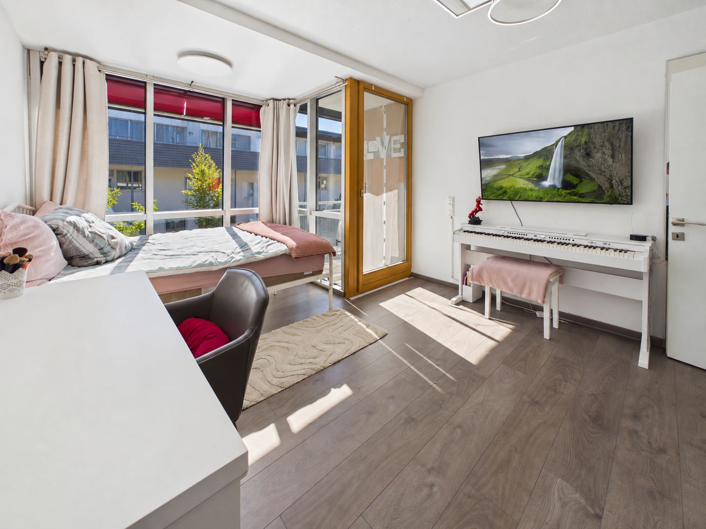 Helles Schlafzimmer mit Bett, Schreibtisch, Sessel, großem Fenster und Klavier an der Wand.
