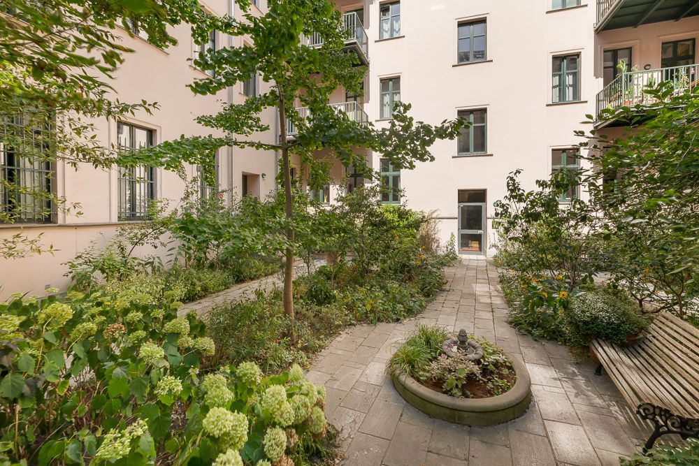 Begrünter Innenhof mit Bäumen, Blumenbeeten, Gehweg und Holzbank, umgeben von Wohngebäuden.