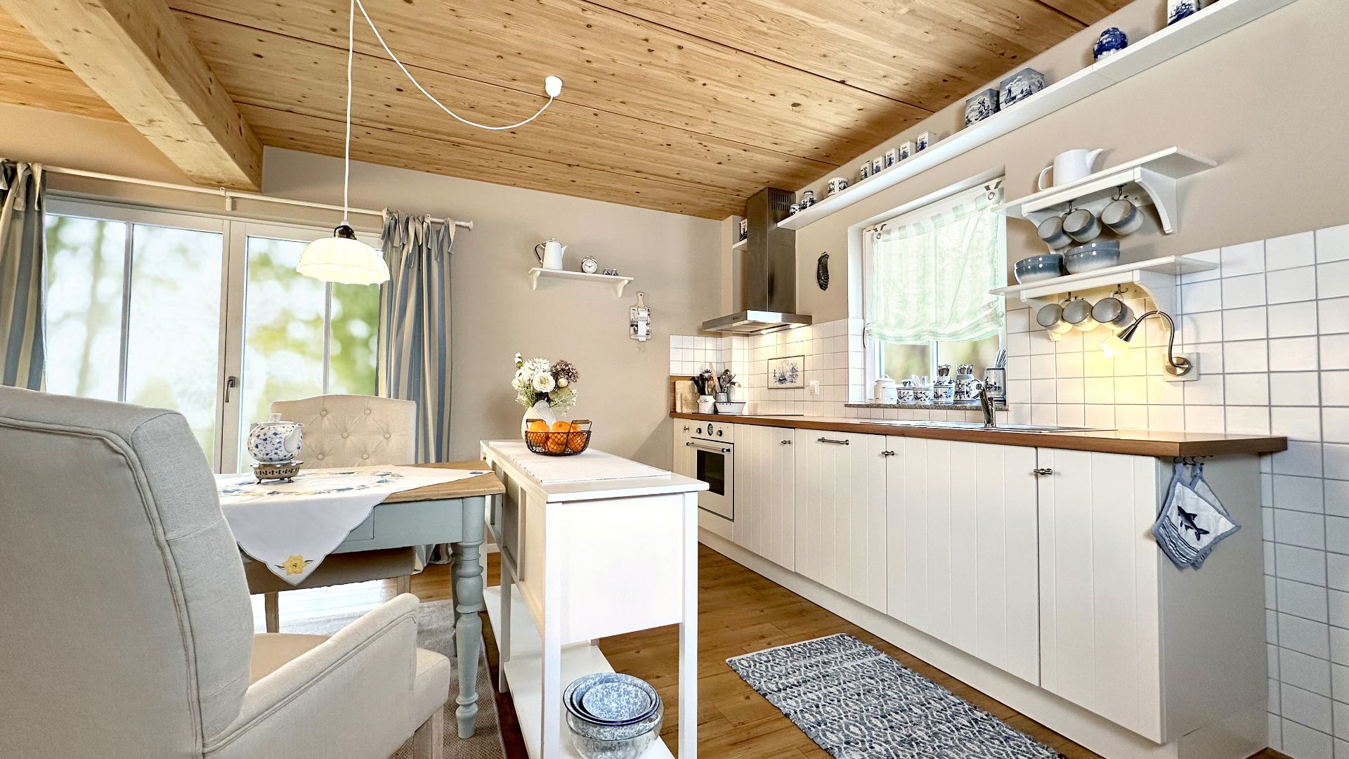 Helle Wohnküche mit Holzdecke, Esstisch, weißen Schränken und Fenstern mit Vorhängen.