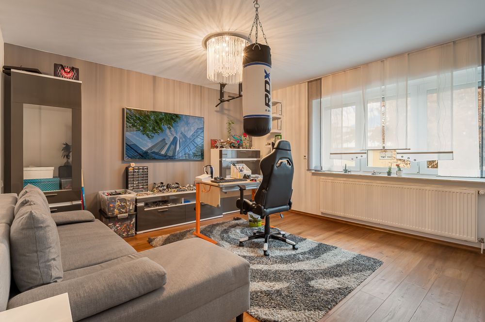Modernes Wohnzimmer mit grauem Sofa, Schreibtisch, Gaming-Stuhl und Boxsack, großes Fenster mit Gardinen.