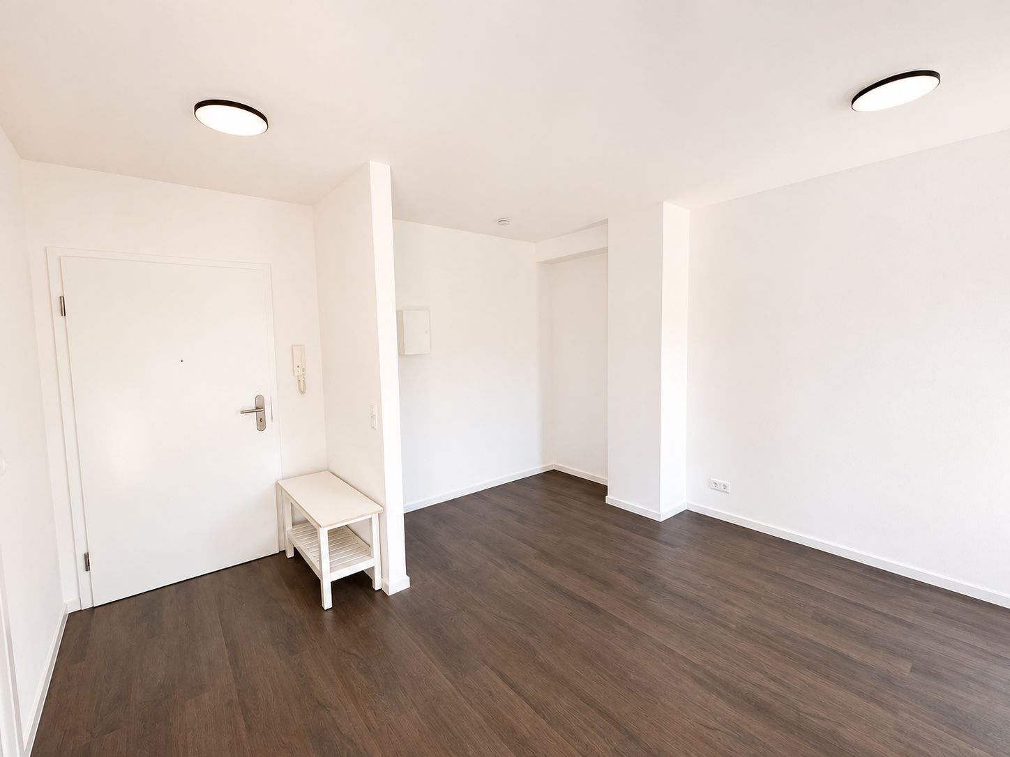 Leerer, heller Raum mit dunklem Holzboden, weißen Wänden und kleiner weißer Bank.