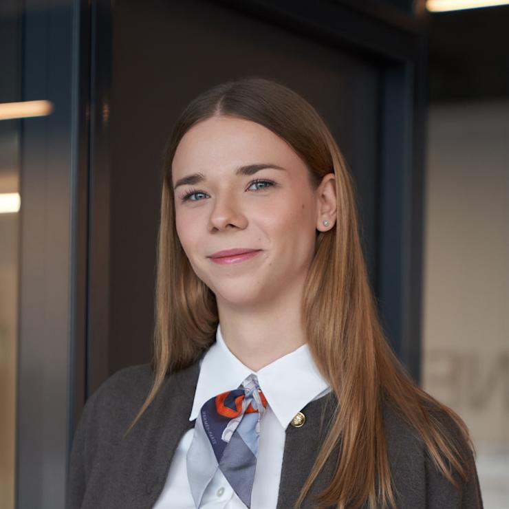 Junge Frau mit langem Haar, weißer Bluse und Halstuch steht in einem modernen Innenraum.