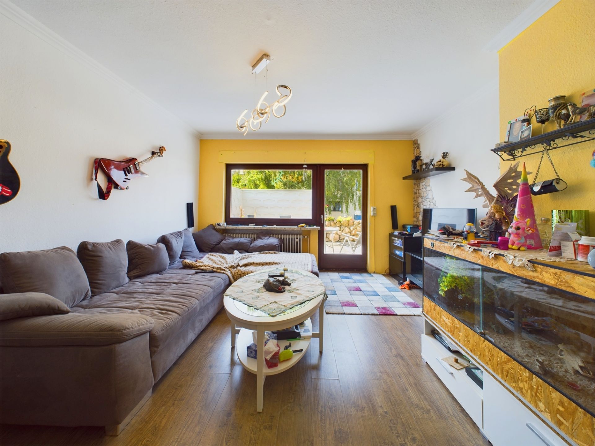 Wohnzimmer mit grauer Eckcouch, Aquarium, Wandregalen und Glastür zum Garten.