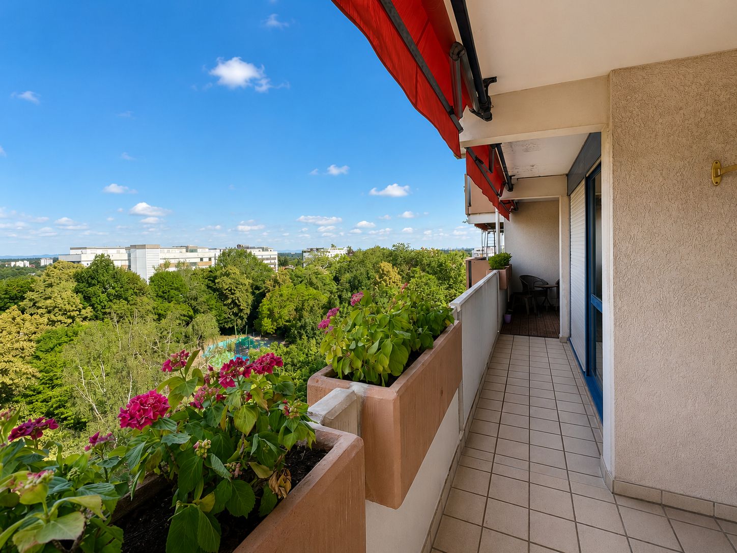 Langer Balkon mit roten Markisen, Blumen und Blick auf grüne Bäume und Gebäude.