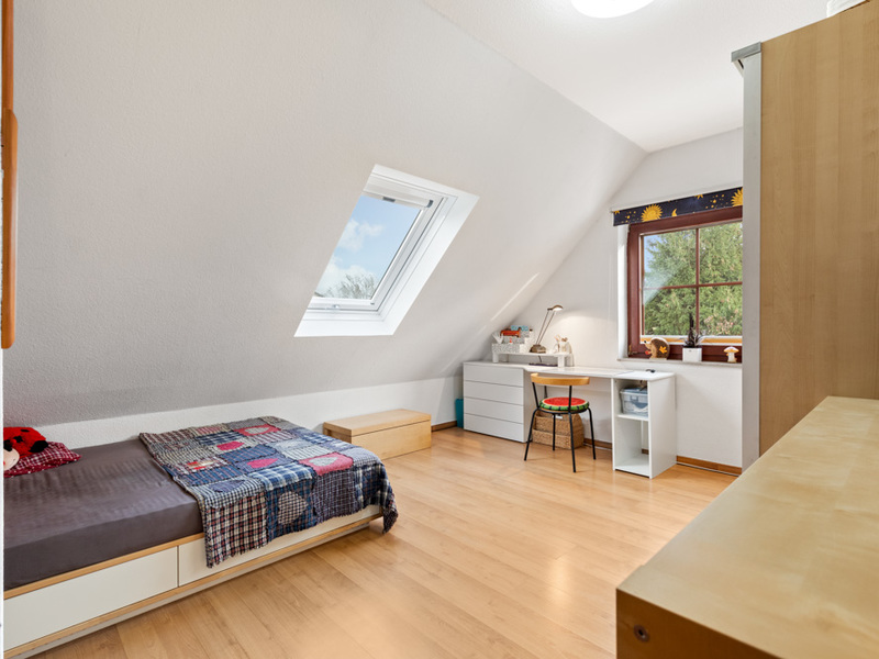Helles Zimmer mit schräger Decke, Einzelbett, Schreibtisch am Fenster und Holzfußboden.