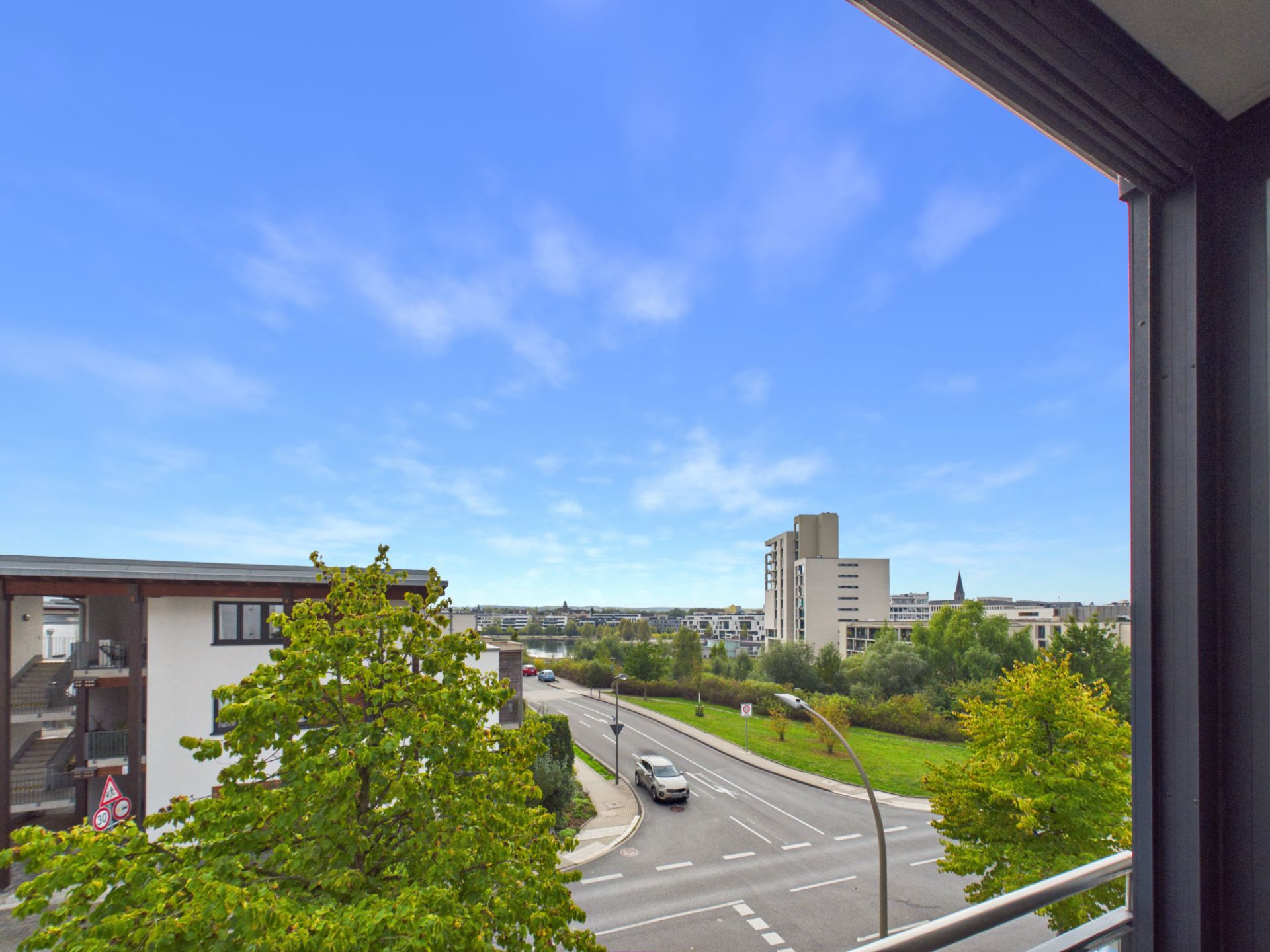 Blick von einem Balkon auf eine Straße, Bäume und mehrere Wohngebäude bei klarem Himmel.