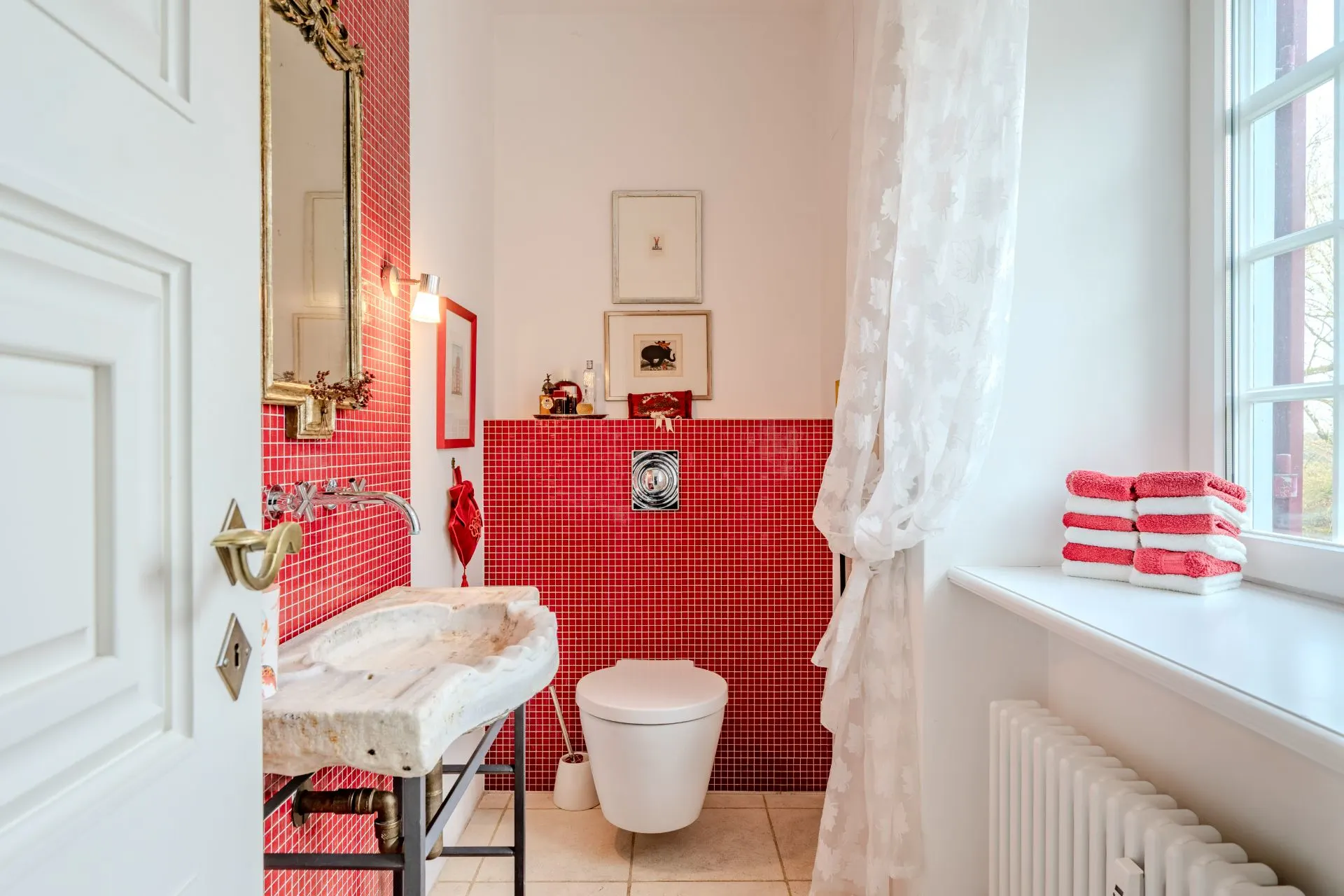 Helles Bad mit rotem Fliesenspiegel, Waschbecken, WC und Fenster mit weißen Vorhängen.