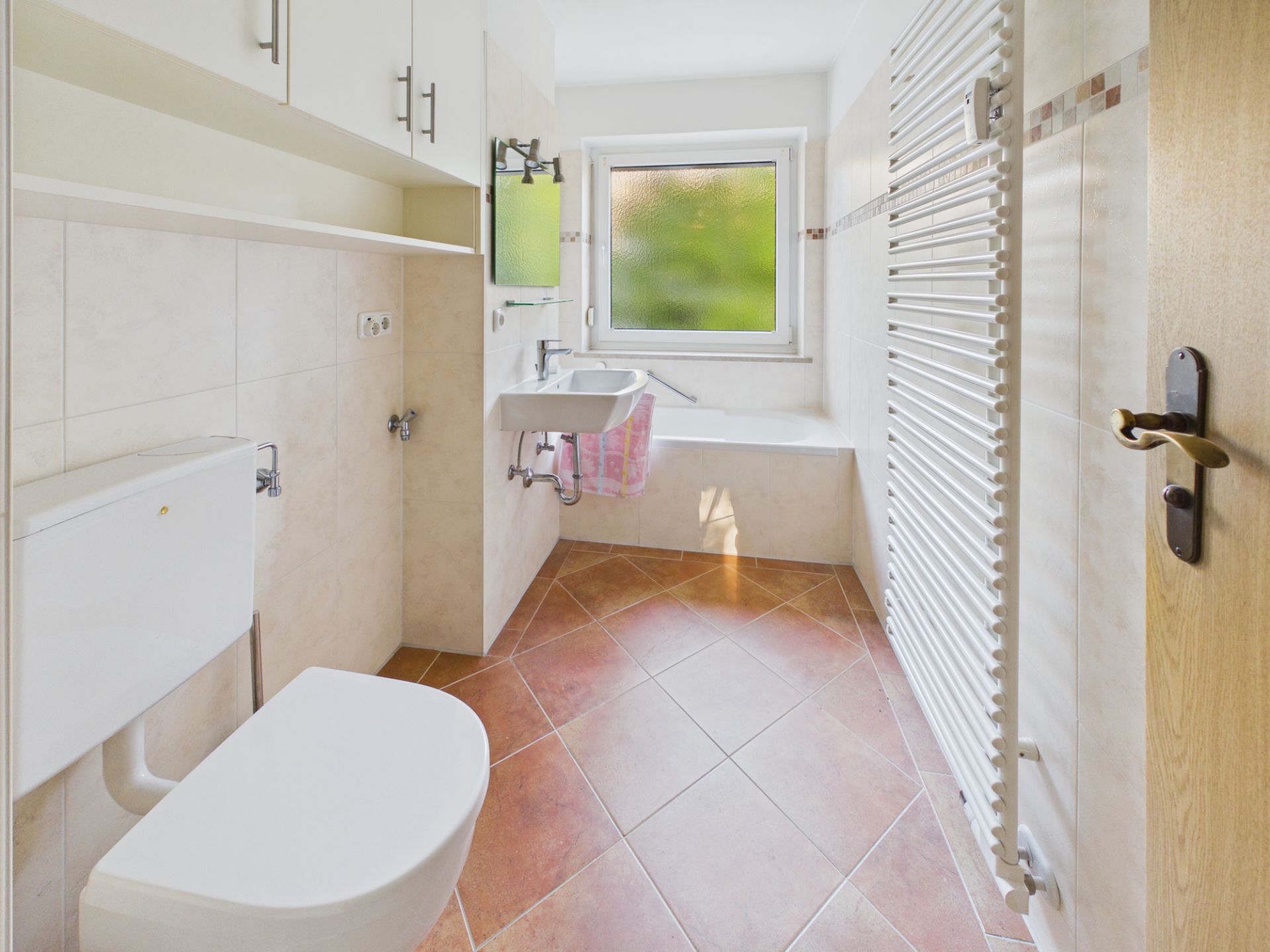 Helles Badezimmer mit Toilette, Waschbecken, Badewanne, Heizkörper und Fenster.