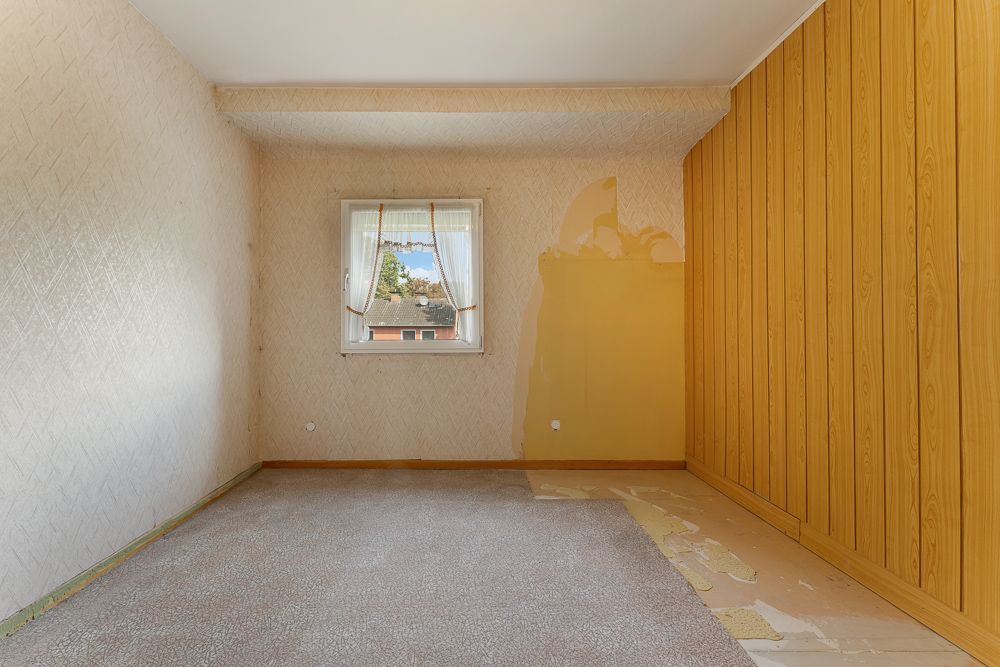 Leeres Zimmer mit Teppichboden, einem Fenster und einer Holzvertäfelung an einer Wand.
